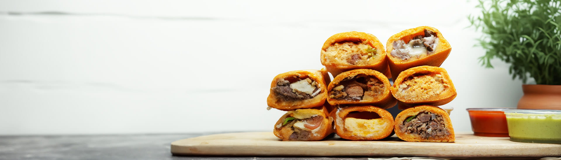 various empanadas stacked on a wooden board besides sauces